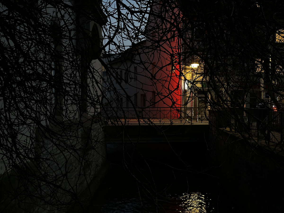 Ein beleuchteter Übergang über einen Wasserlauf, umgeben von dunklen Ästen und Gebäuden.