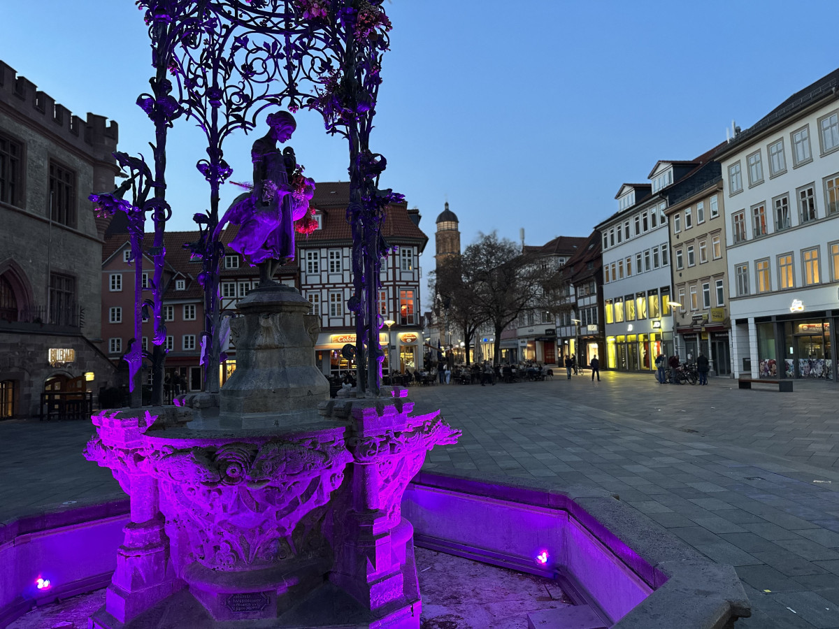 Gänseliesel mit Blick auf St. Albaniin einem Platz, umgeben von historischen Gebäuden und Abendbeleuchtung.